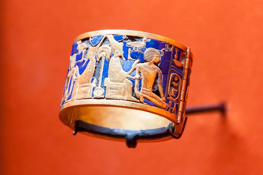 The bracelet of Queen Ahhotep, with Geb on his throne: A hinged gold bracelet inlaid with lapis lazuli, bearing the name of King Ahmose I. One half shows the earth god Geb, wearing the double crown, enthroned with his hands on the shoulder of the kneeling king.